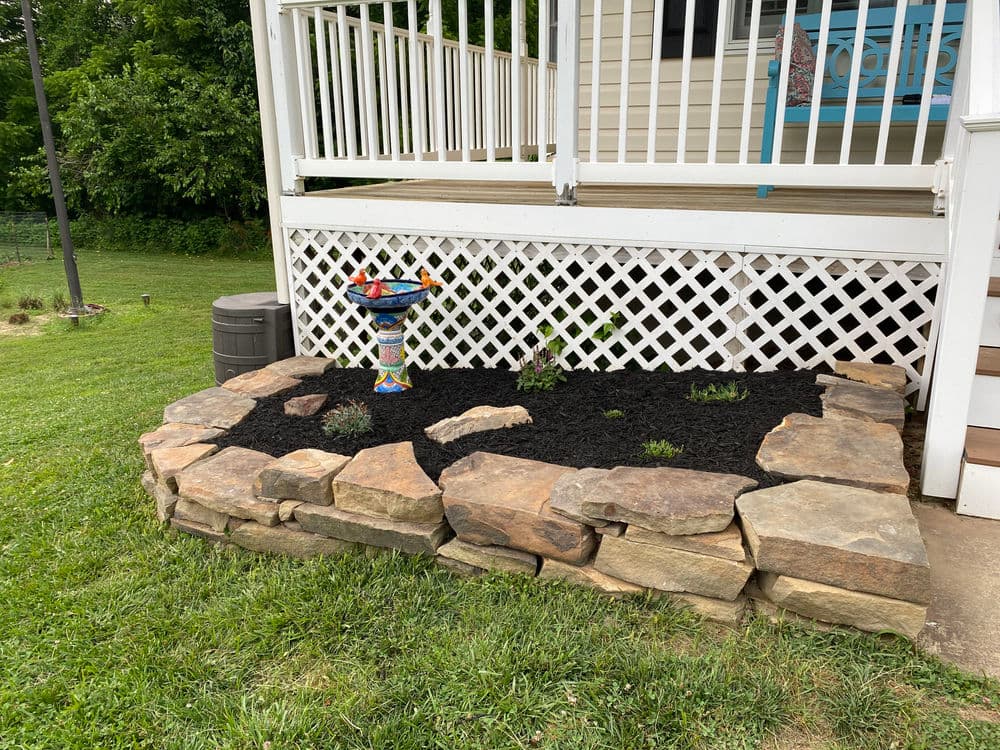 Decorative stone garden bed with black mulch and colorful birdbath under a porch.