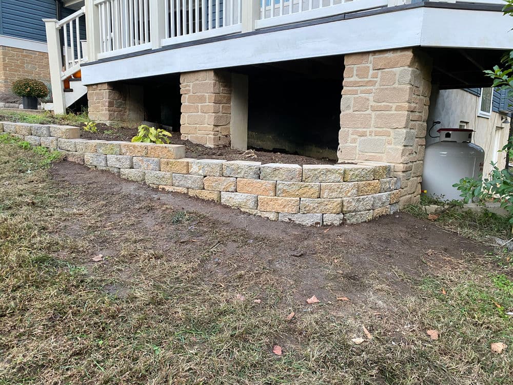 Stone retaining wall built under a deck, with freshly landscaped area and propane tank in view.