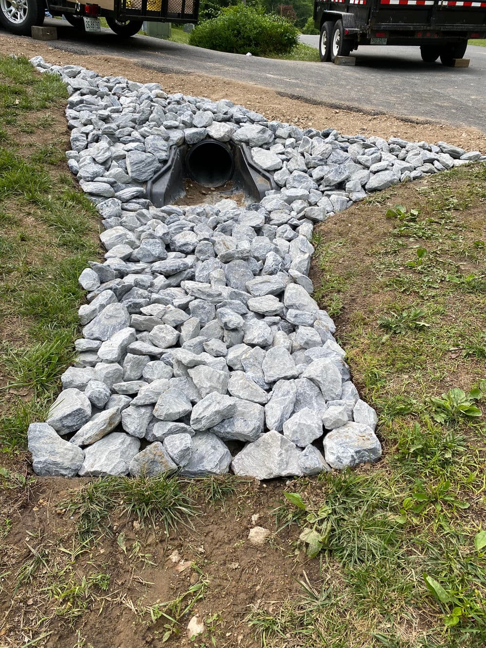 Rock drainage installation leading to a culvert, surrounded by grass and gravel.