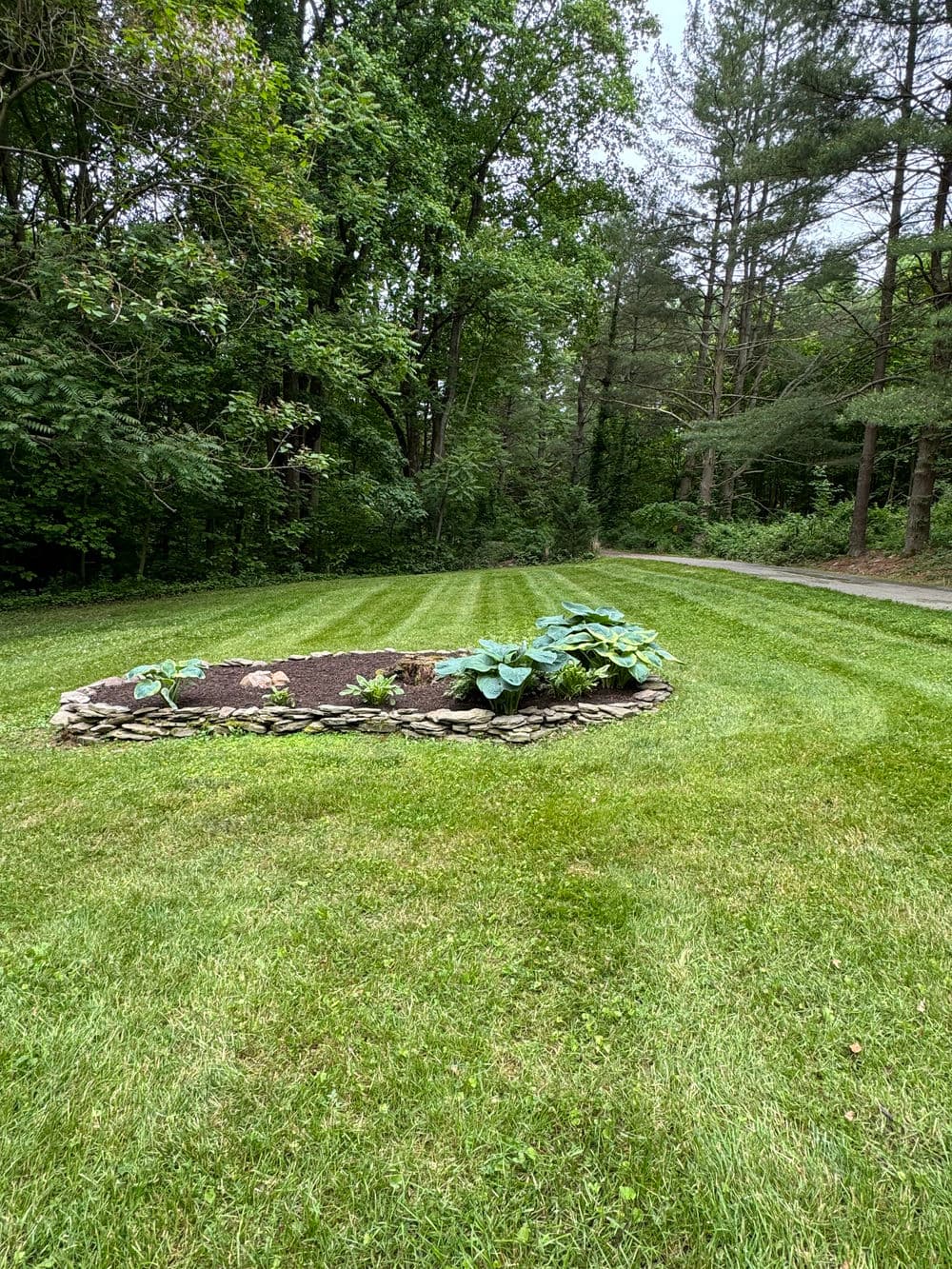 Lush green lawn with a circular stone garden bed featuring various plants and trees in background.